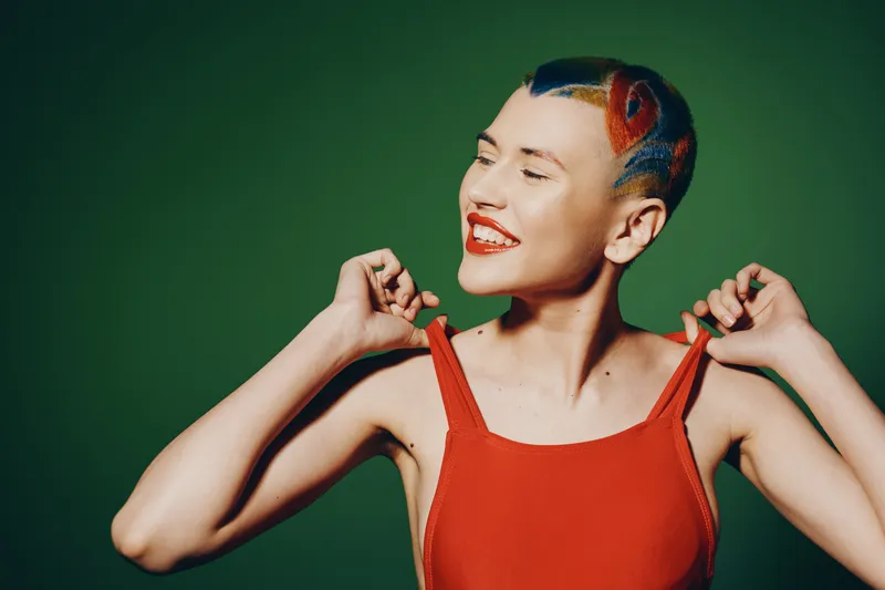 young woman with creative shaved hairstyle wearing red tank top smiling joyfully against green background, expressing happiness and vibrant personality