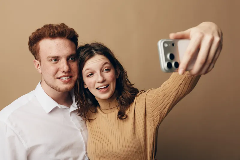 Young couple taking selfie with smartphone, smiling and posing together against beige background. Casual clothing, happy mood, modern technology, social media concept, close-up portrait.