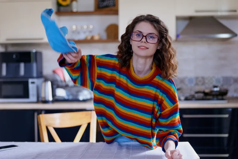 Cheerful woman indoors, cleaning a bright kitchen, wearing a colorful striped sweater, wiping a surface with a cloth, relaxed posture, warm natural light, modern home decor backdrop