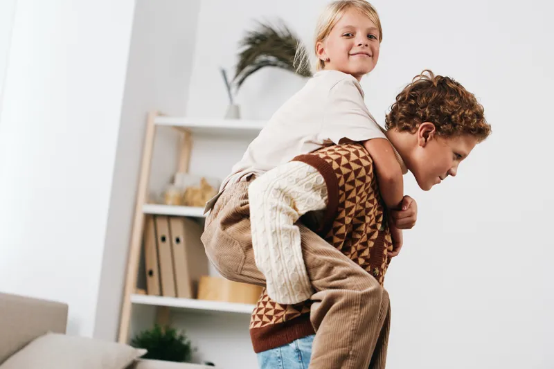 Two playful children share a piggyback moment indoors, smiling as they balance on each other’s backs in a cozy, sunlit room filled with warm home ambiance.