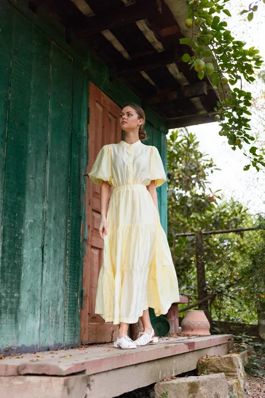 Woman in a pale yellow dress stands on a rustic porch beside a green wooden wall, basking in soft outdoor light with leafy surroundings and a serene countryside vibe.