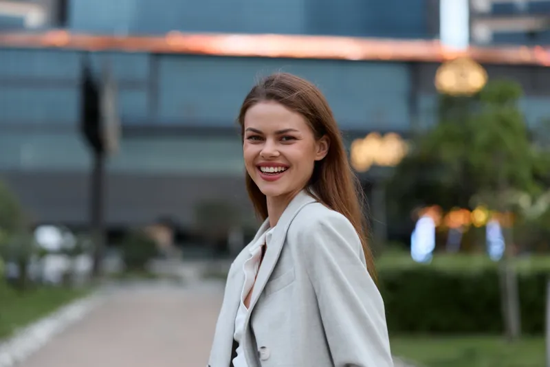 Happy business woman smiling outdoor in light gray blazer with blurred urban background and natural green elements in spring afternoon light
