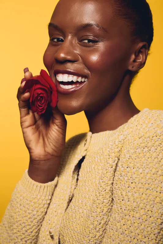 Smiling black woman with dark skin holding red rose near face wears yellow knitted sweater on yellow background. Concept of beauty, skincare, happiness, and natural glow in studio portrait.