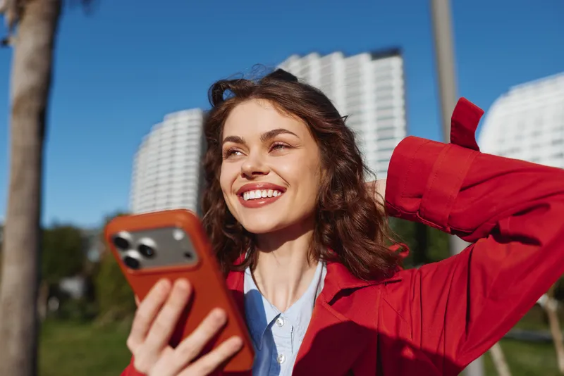 Smiling woman in a red jacket takes a selfie with a smartphone outdoors, urban backdrop of tall buildings, sunny day, casual fashion, cheerful mood.