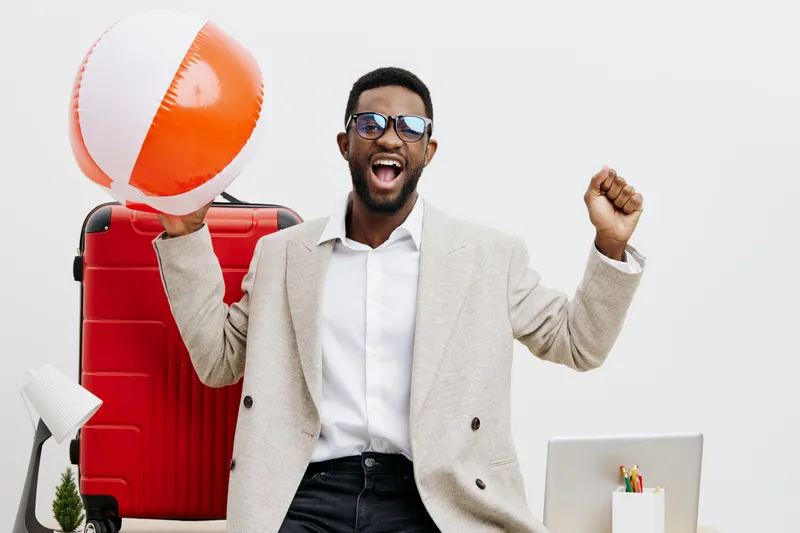 Smiling young man with dark skin wearing sunglasses, beige blazer, and white shirt holding a beach ball in a bright travel-themed studio setting. Summer vacation, happiness, leisure, lifestyle, travel