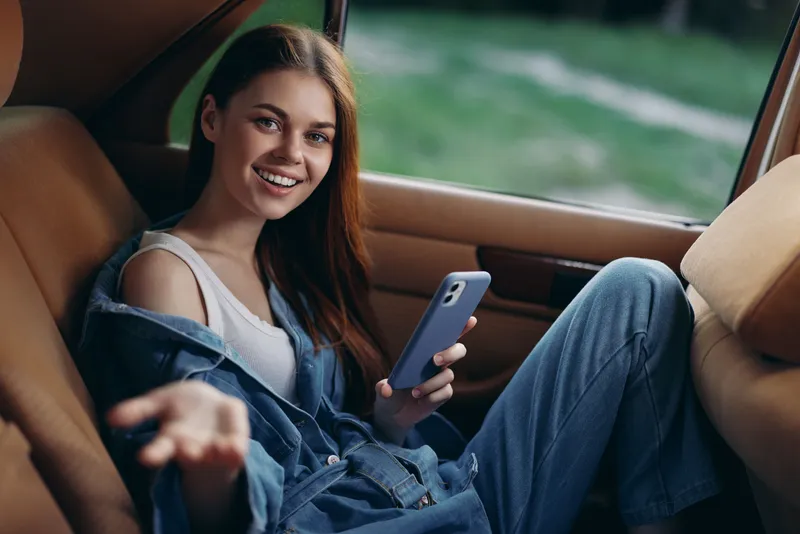 Smiling young woman with long hair in casual outfit enjoying time in a car while using smartphone, ideal for lifestyle and technology themes.