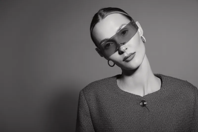 Portrait of a young Caucasian woman with pale skin wearing large sunglasses and textured jacket against plain background.