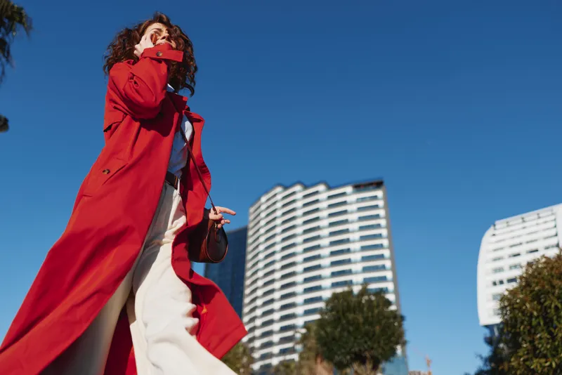 A woman in a red coat walks confidently through an urban landscape under a clear blue sky, highlighting street style, movement, and modern city fashion.