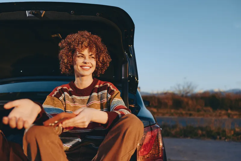 Woman lifestyle in car, she sits on the seat and smiles, curly hair visible at the window, traveling on rural road at sunset, outdoors countryside landscape, warm evening light