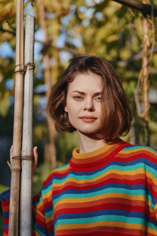 Woman portrait in colorful striped sweater leaning on bamboo pole in outdoor garden, young adult with bob haircut and calm expression, relaxed lifestyle fashion headshot with natural light.