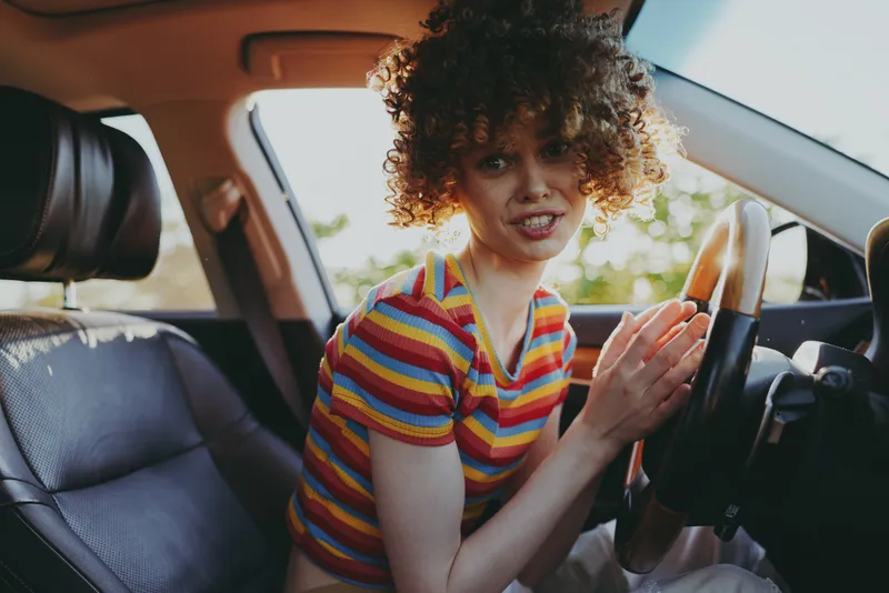 Cheerful woman in a car, colorful striped shirt, sunlit interior, retro film color, candid lifestyle moment, relaxed posture, warm tones, natural light, subtle smile, outdoor blur