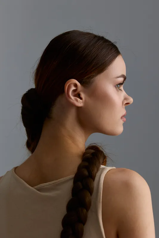 Profile of young woman with long braided hair and light beige top, looking thoughtful against a simple gray studio background.