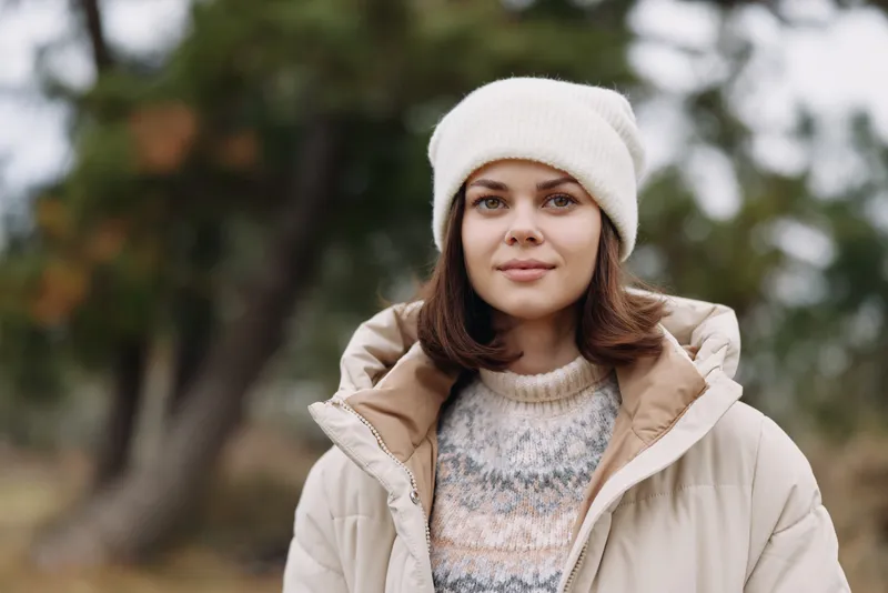 Woman in a warm coat and knit sweater, outdoors in a wooded setting, calm portrait with soft winter light and cozy fashion atmosphere.
