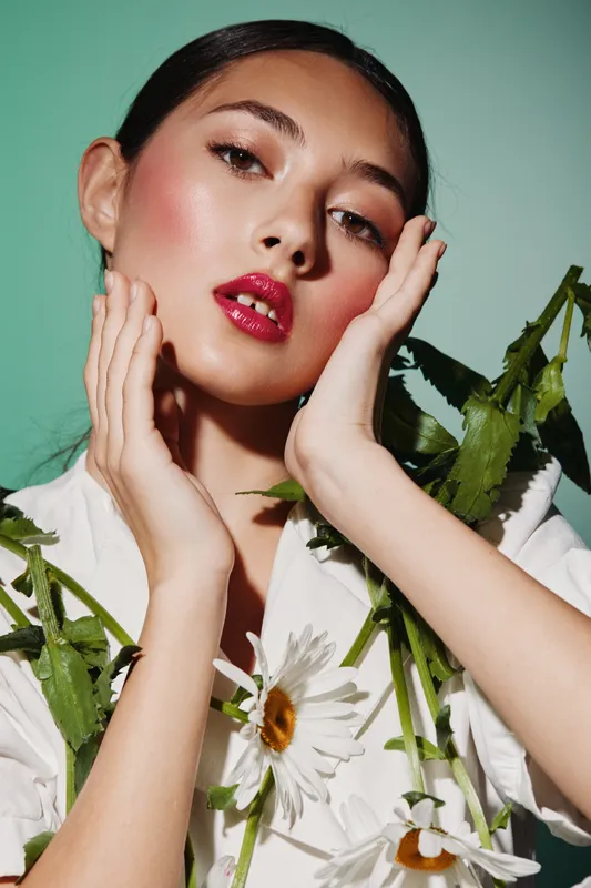 Young woman with glossy pink lips and dramatic eye makeup poses elegantly holding white daisies, wearing stylish white clothing against soft green background.