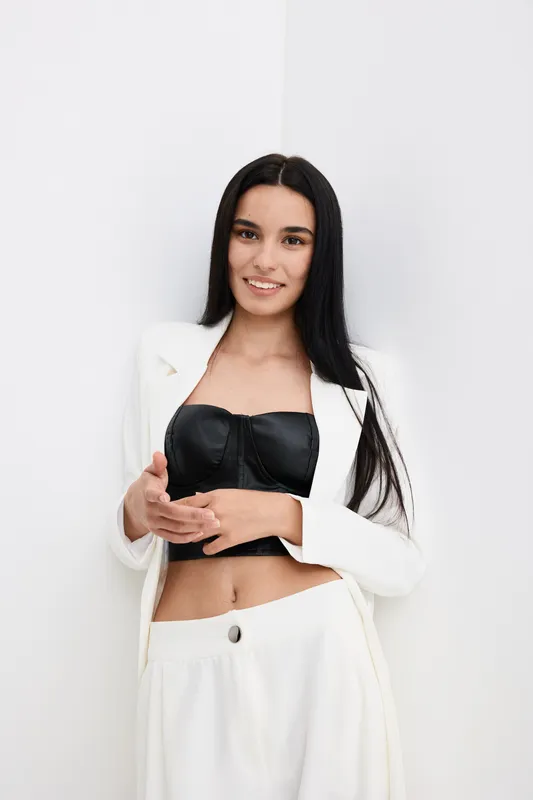 young woman in white suit wearing black crop top, standing near white wall with long dark hair and calm smile, modern fashion style portrait indoors
