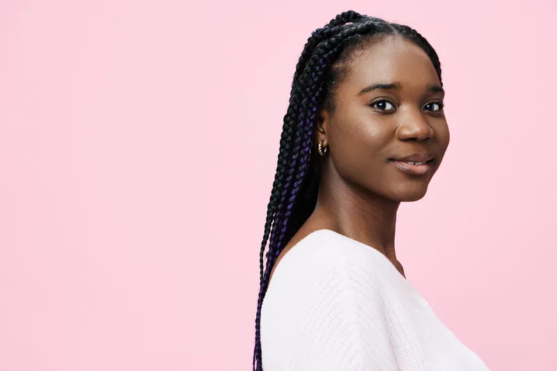African woman with braided hair smiling against a pink background, showcasing beauty and confidence in a stylish white sweater