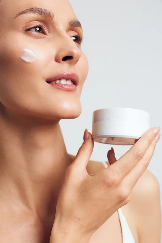 Young woman with clear skin holds white jar of cream applies skincare product on cheek smiling with closed lips isolated on light background beauty routine concept.