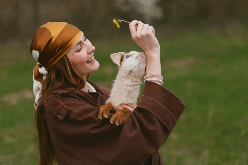 Smiling woman in a brown cloak embraces a playful goat kid outdoors, a warm lifestyle moment with nature, affection, and gentle light creating a cozy, intimate mood.