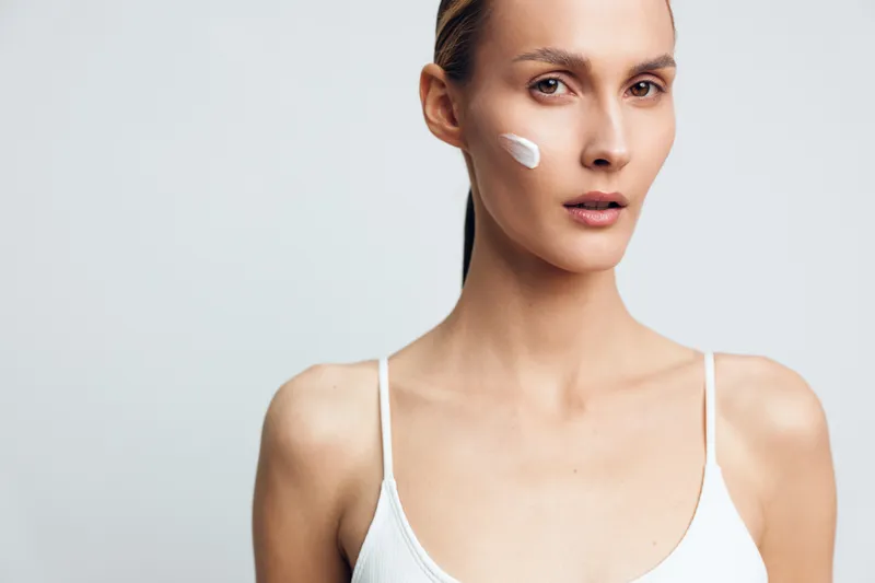 Young woman with clear skin applying white cream on cheek, natural beauty concept, minimal makeup, soft lighting, simple background, skincare routine, healthy complexion