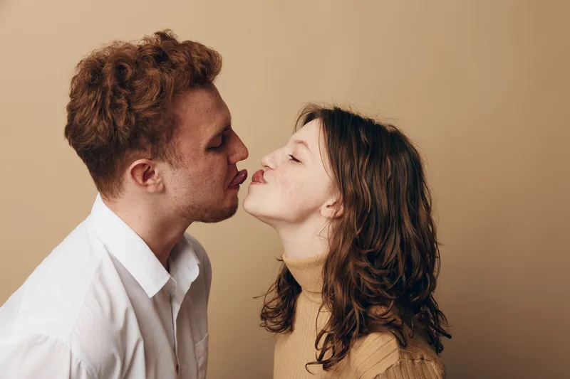 Young couple with red hair and brown hair leans in for a kiss on beige background. Intimate moment, love, affection, and romantic connection between man and woman.