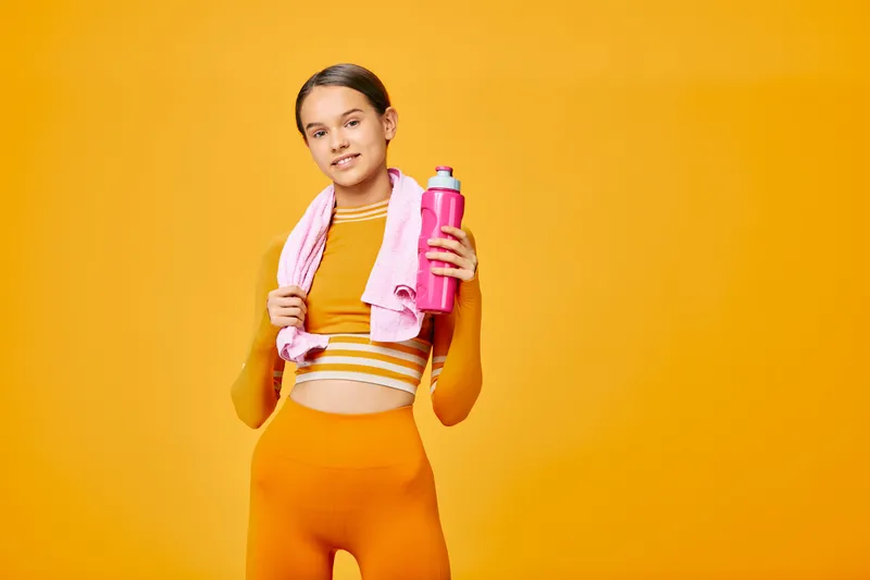 Athletic woman in bright orange activewear stands against a vibrant orange backdrop, holding a pink bottle with a towel over her shoulders, ready for a dynamic workout and fitness routine.