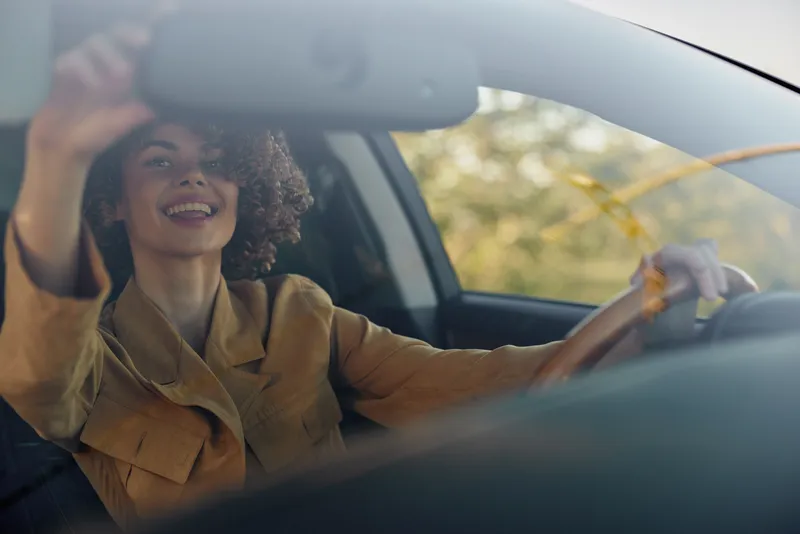 A cheerful woman driver in retro style sits behind the wheel, smiling as sunlight fills the car. Lifestyle portrait conveying casual travel, optimism, and everyday moments.