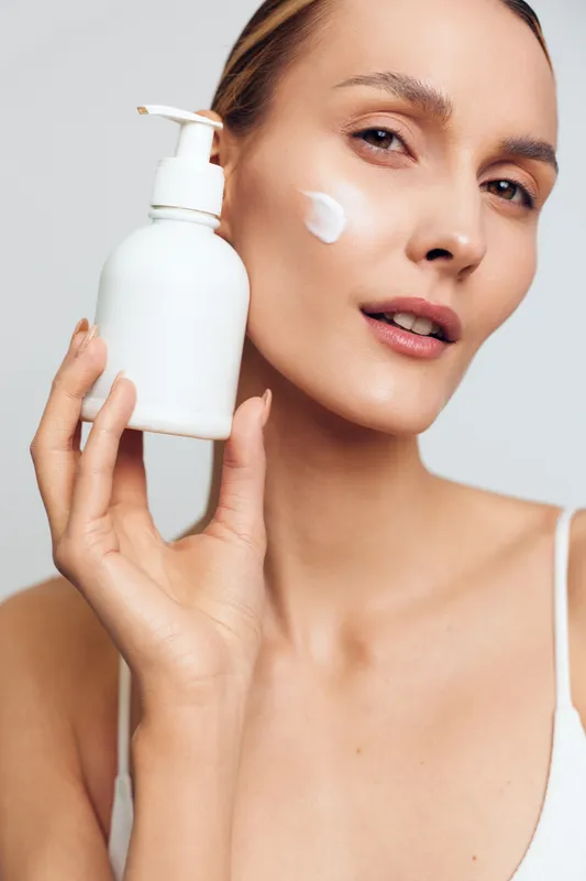 Young woman with brunette hair applies facial cream from a white bottle in a close up skincare moment, conveying calm focus, soft lighting, and smooth skin