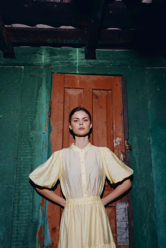 Stylish portrait of a woman in a pale yellow dress standing with hands on hips in front of a green doorway, creating a bold artistic fashion vibe.