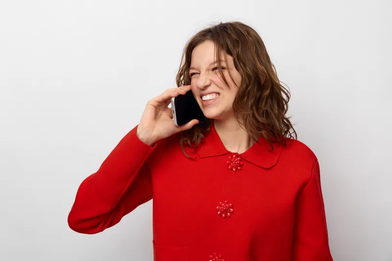 Young woman in red sweater talking on smartphone with frustrated expression. Isolated on white background. Concept of communication, stress, and emotional reaction during phone call.