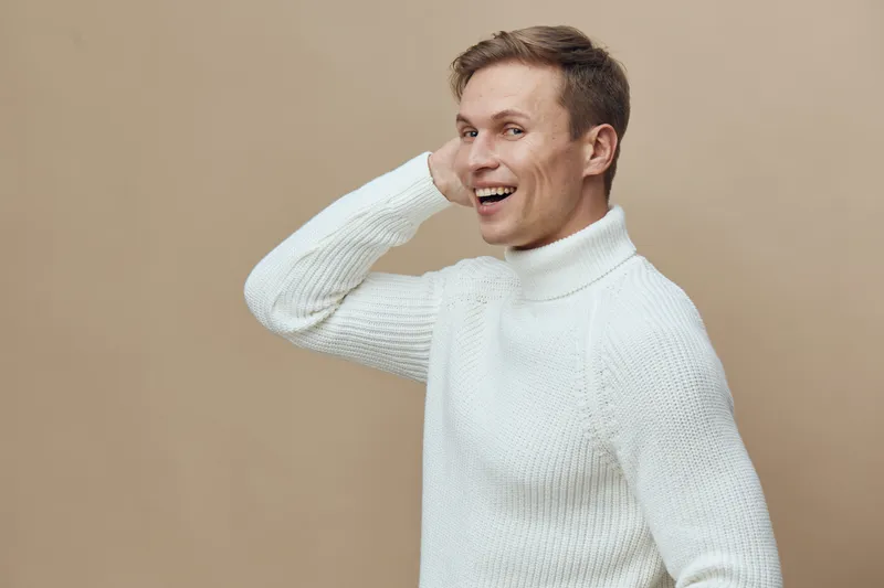Happy smiling young man with short hair wearing white sweater, posing against beige background. Joyful, cheerful, casual, fashion, portrait, studio shot, lifestyle concept, modern clothing.