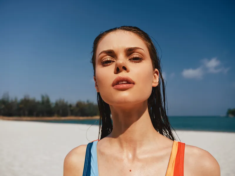 Close up portrait of a young woman at a sunny beach, wet hair, sunlit skin, bold gaze, and colorful swimsuit straps, with blue sky and calm sea behind.