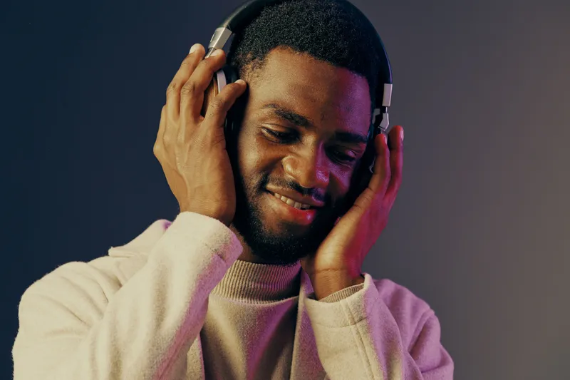 Young Black man enjoying music with headphones, smiling joyfully in cozy sweater against colorful background, concept of relaxation and happiness