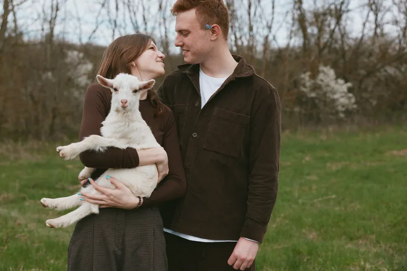 Young couple embraces outdoors while woman holds a white baby goat in her arms during a relaxed lifestyle moment in early spring nature.