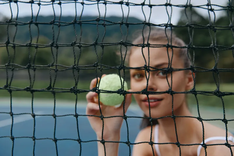 Female athlete holding a tennis ball against the net, smiling confidently on a sunny day, showcasing sports spirit and determination on the court