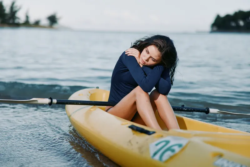 A lone woman sits in a yellow kayak on calm water, hugging her knees with the paddle nearby, conveying solitude, focus, and quiet coastal reflection.