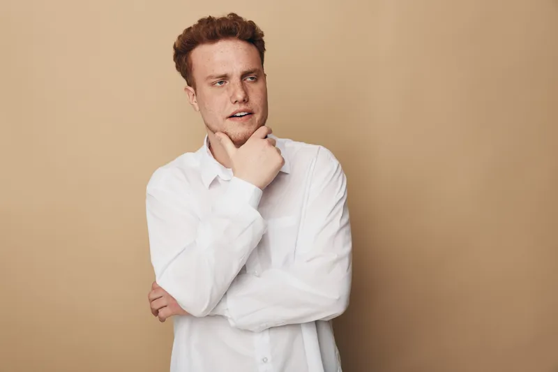 Thoughtful young man with red hair and beard wearing white shirt looks away with hand on chin. Neutral beige background. Concept of contemplation, decision making, and introspection.