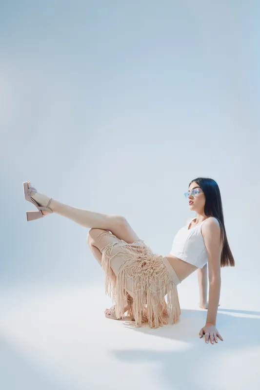 Young woman in stylish beige fringed skirt and white top poses sitting on floor with legs raised. Clean background highlights beauty and fashion concept.