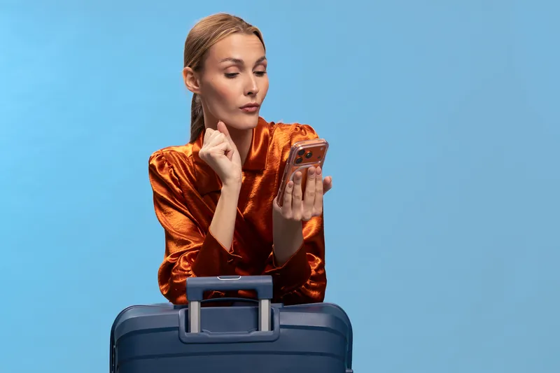 Young woman in orange shirt looks thoughtfully at smartphone while leaning on blue suitcase. Travel concept with focused female traveler, blue background, and casual outfit for trip preparation.