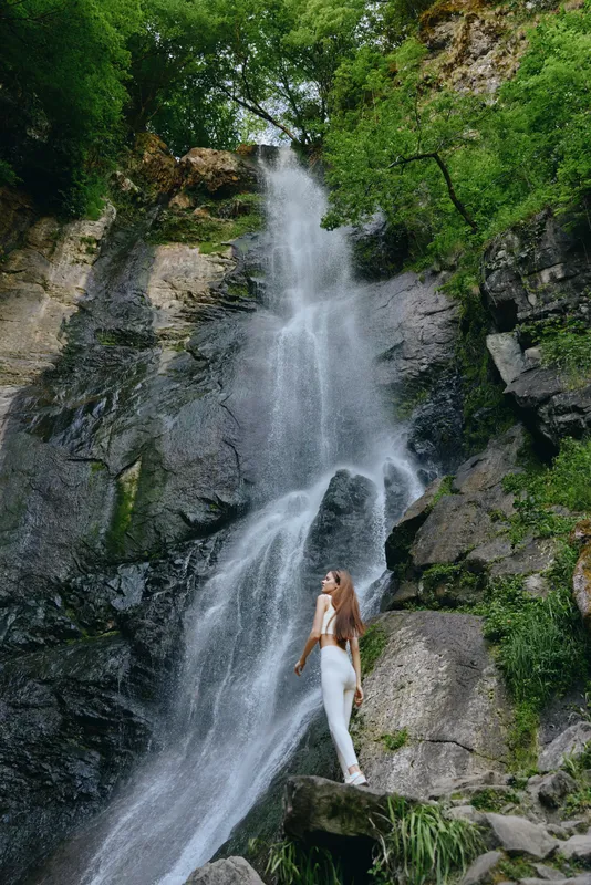 Woman exploring waterfall nature scene, wearing white outfit, surrounded by lush green forest and rocky cliffs under natural daylight.