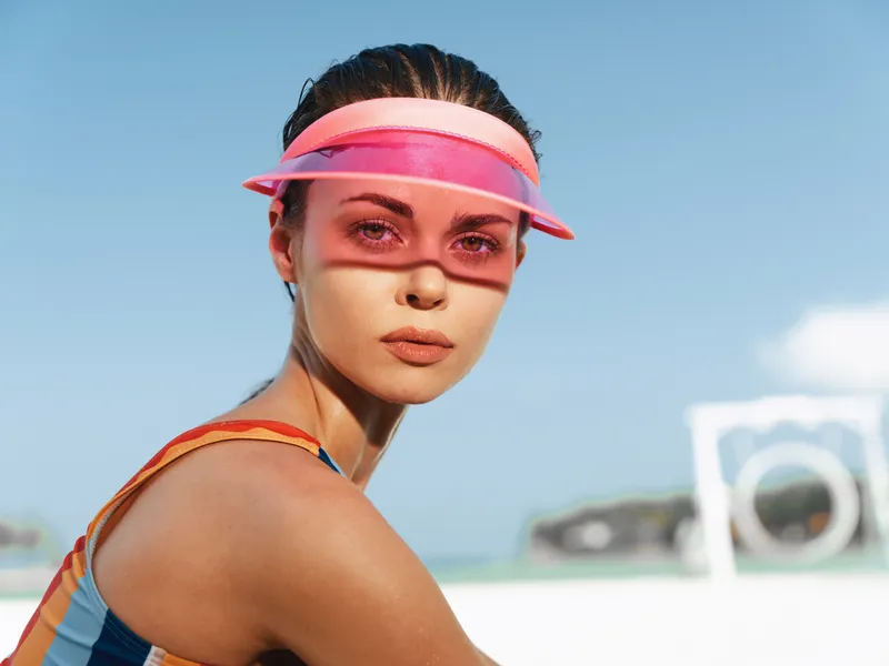 Portrait of a youthful woman on a sunny beach wearing a pink visor and colorful athletic top. Close framing highlights smooth skin, confident gaze, and summer mood.
