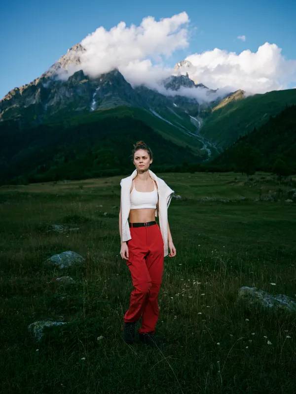 Mountain landscape with a grassy meadow, cloudy sky, and a confident woman in a white crop top, redpants and a light jacket, showcasing bold outdoor style and adventure