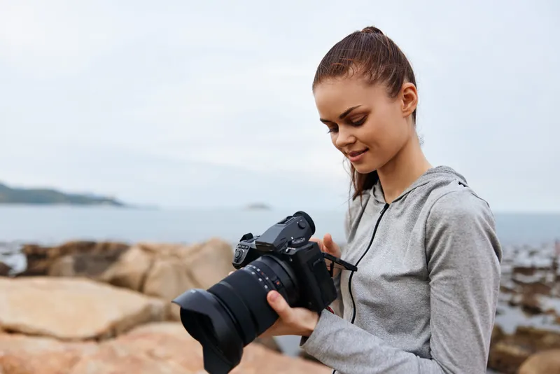 Young woman in casual hoodie enjoying photography by the sea, showcasing passion for capturing moments with a camera in a serene outdoor setting