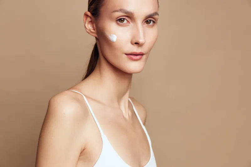 Young woman with clear skin and natural makeup applying cream on her face, wearing white top, neutral background, skincare routine, calm expression, soft lighting.