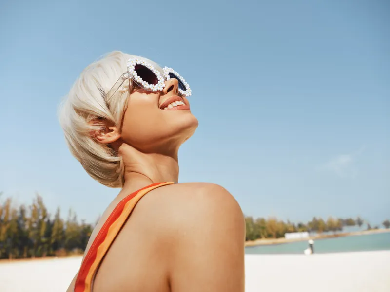 Portrait of a smiling woman on a sunny beach, wearing stylish white sunglasses and an orange swimsuit strap, bathed in bright daylight against a clear blue sky.