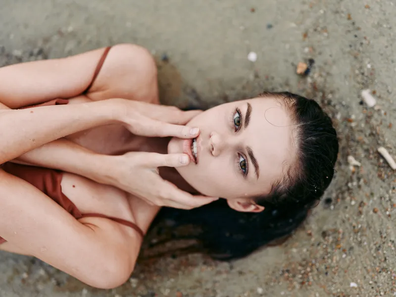 Closeup portrait of a young woman lying on a sandy beach, head tilted, hands framing her face, serene gaze, sunlit skin, wind in hair, intimate and dreamy.