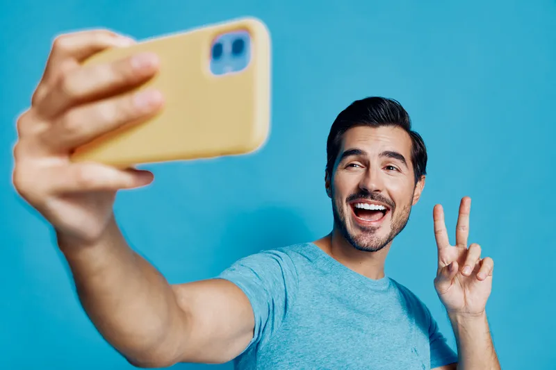 Young smiling man taking selfie with mobile phone against a bright blue background, showcasing a fun and cheerful personality in a relaxed setting.