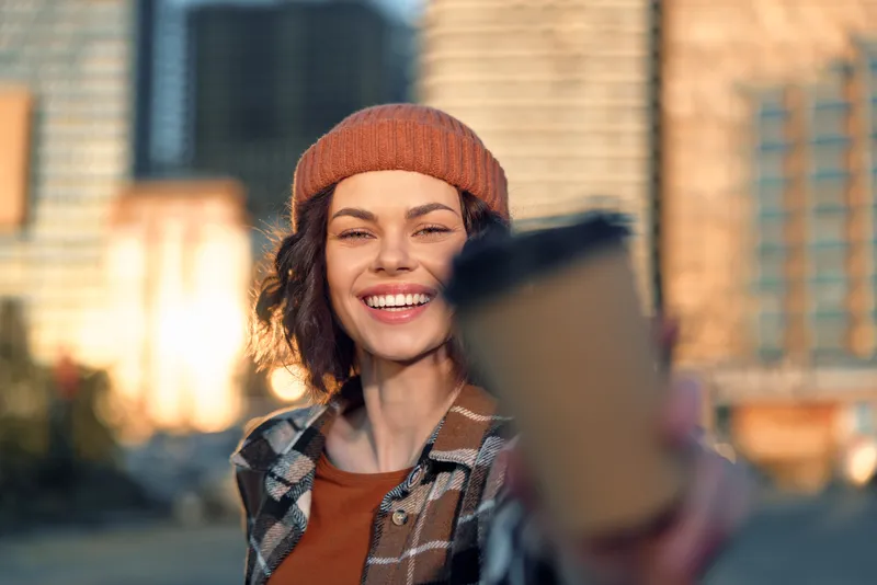 Woman smile coffee city portrait beanie sunlight urban young woman extends takeaway mug in street with golden hour glow, authenticity and candid lifestyle captured in a mindful living moment.