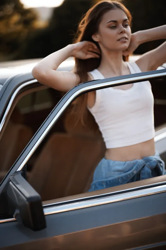 Young woman enjoying summer vibes, wearing a white tank top and denim shorts, leaning against a classic car with a relaxed expression in a natural outdoor setting.