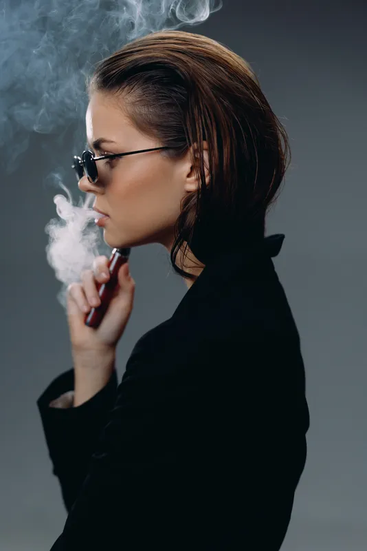 Stylish woman vaping indoors with smoke around, wearing sunglasses and black outfit, modern and moody atmosphere