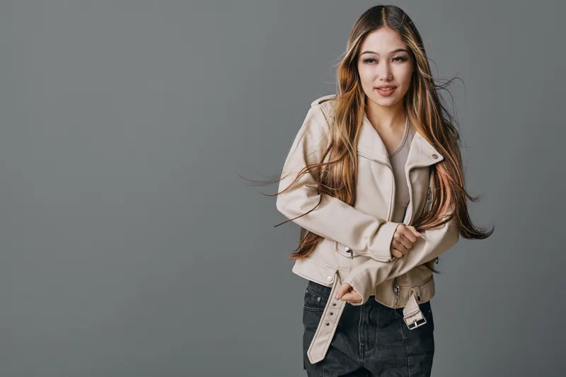 Young woman with long hair wearing beige jacket and black jeans posing on gray background looking away. Casual fashion style with natural makeup and relaxed expression outdoors.